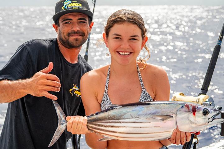Two people on a boat, one holding a fish and both smiling with an ocean background.