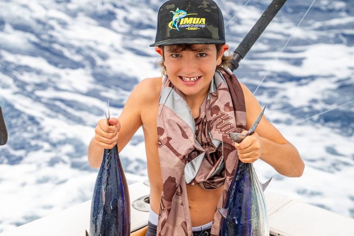 Young person smiling, holding two fish on a boat with ocean in background.