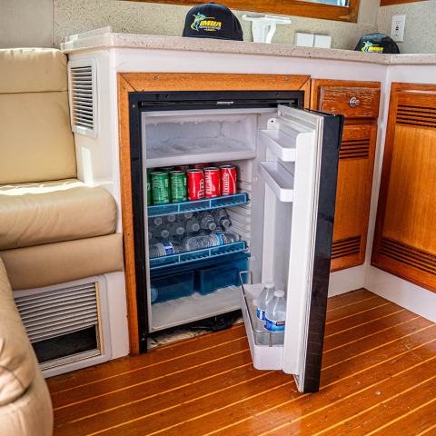 Small fridge open in a boat's cabin, stocked with drinks next to a beige sofa and wooden cabinets.
